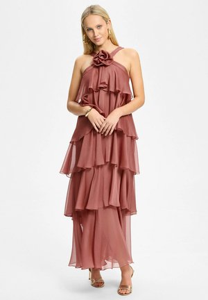 Femme portant une robe à plusieurs couches couleur rose poudré avec un détail floral, boucles d'oreilles anneaux dorés, bracelet et talons ouverts dorés, debout sur fond blanc.