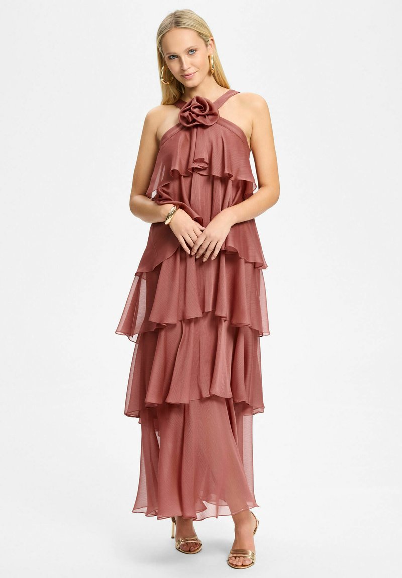 Femme portant une robe à plusieurs couches couleur rose poudré avec un détail floral, boucles d'oreilles anneaux dorés, bracelet et talons ouverts dorés, debout sur fond blanc.