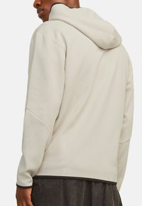 Sweatshirt beige clair à capuche avec une coupe décontractée, présentant des accents noirs le long de la capuche et de l'ourlet. Tissu doux et texturé avec des détails de couture.