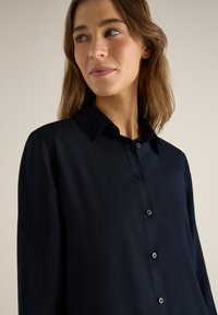 Jeune femme aux cheveux châtain clair portant une chemise noire à col boutonnée, regardant légèrement sur le côté sur un fond uni.