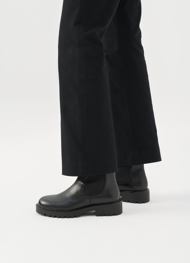 Sorte læder ankelstøvler med strækpaneler på siderne og robuste gummisåler, sat sammen med brede sorte bukser mod en neutral baggrund.