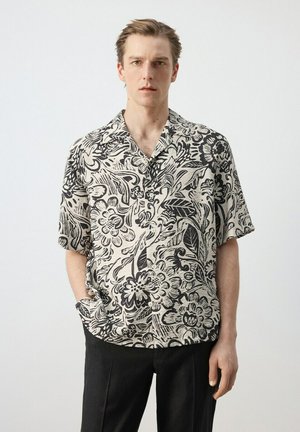 Joven con camisa de manga corta de estampado floral en blanco y negro y pantalones negros, de pie frente a un fondo claro y liso.