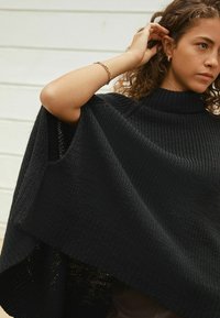 Woman adjusting curly hair wearing black knitted poncho with turtleneck and gold hoop earring and bracelet against white background.