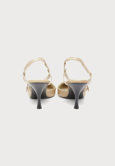 Escarpins beige à bout ouvert, à bride arrière, avec fines brides réglables et talons aiguilles noirs sur fond blanc uni.