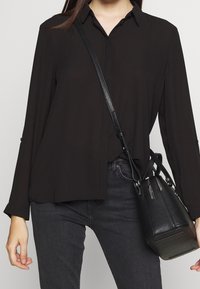 Camisa negra de manga larga con botones y un corte relajado, combinada con jeans oscuros y un pequeño bolso cruzado de cuero negro con acentos texturizados.