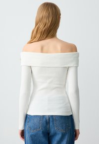 Weiße Off-Shoulder-Bluse mit langen Ärmeln und geripptem Textur, taillierte Silhouette und weitem Ausschnitt, kombiniert mit hellblauen, hoch taillierten Jeans.