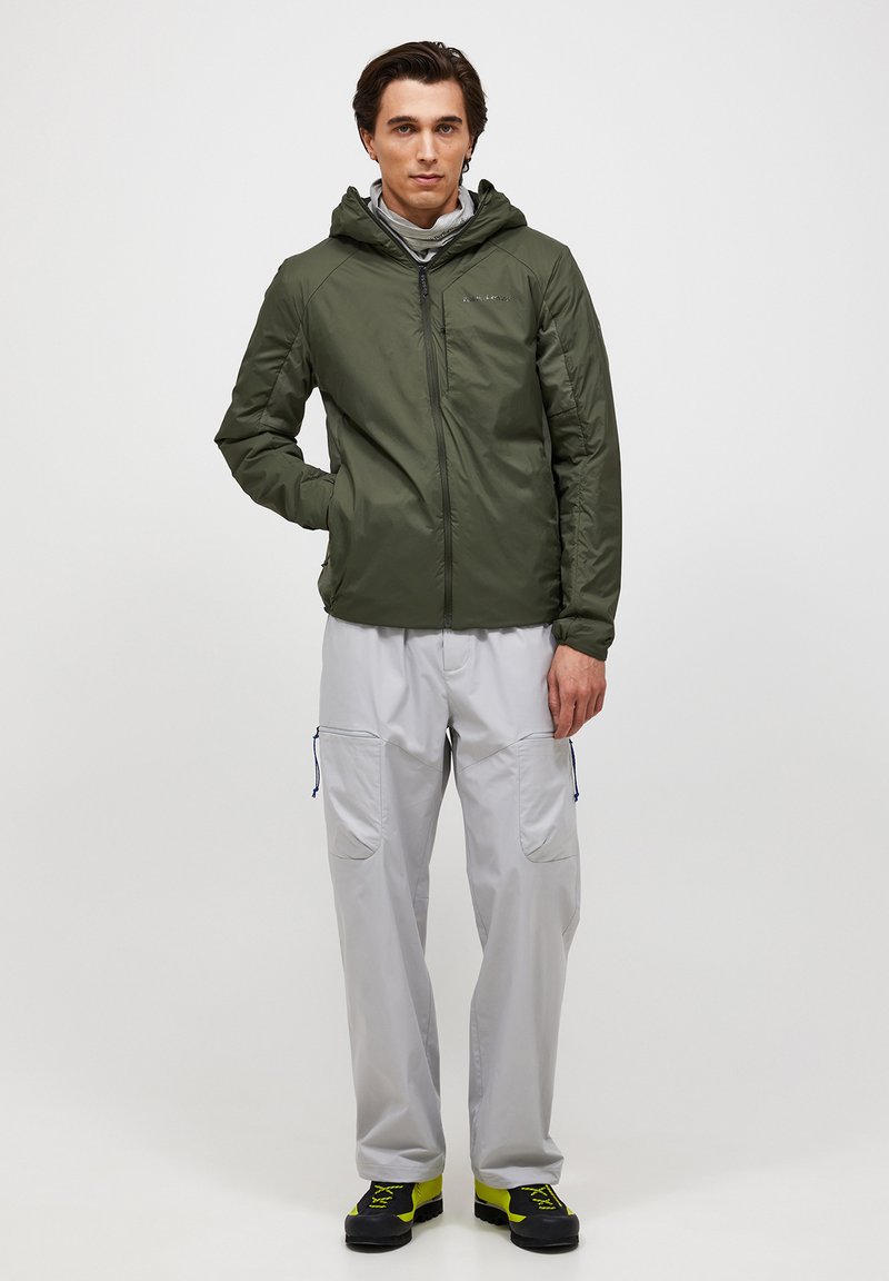 Man draagt een olijfgroene gewatteerde jas met capuchon, lichtgrijze cargobroek en zwarte schoenen met neongele accenten, staat met één hand in de zak.