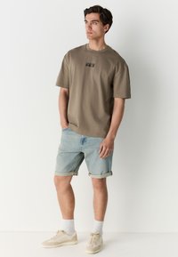 Khaki T-Shirt mit einem Box-Logo, kombiniert mit hochgekrempelten hellblauen Jeansshorts. Die Schuhe sind beige Sneaker, dazu weiße Sportsocken.