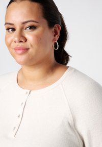 Vrouw met donker haar opgestoken, draagt zilveren creolen en een crèmekleurige blouse met knopen, iets naar links gericht tegen een witte achtergrond.