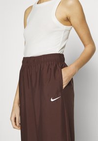 Hnědé sportovní kalhoty s elastickým pasem, bočními kapsami a bílým logem Nike na levé straně. Nosí se s bílým tílkem.