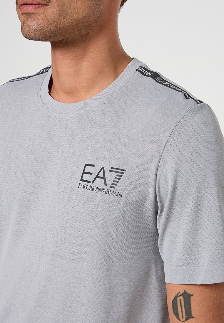Šedé tri čko s krátkým rukávem a kulatým výstřihem, zdobené černým logem EA7 a značkováním na ramenech, vyrobené z texturované látky.