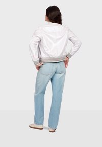 Femme debout, mains sur les hanches, portant une veste bomber en satin blanc, un jean droit bleu clair et des baskets beiges sur un fond uni.