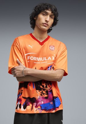 Junger Mann mit lockigem Haar, verschränkten Armen, trägt ein oranges Puma Formula 1 Racing League Trikot mit bunten abstrakten Mustern.