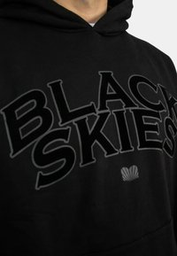 Black hoodie featuring bold, raised text "BLACK SKIES" in a contrasting color, with a shell graphic accent at the bottom. Soft fabric texture.