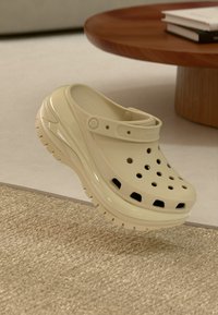 Helle beige Clog mit Belüftungsöffnungen, weicher Textur, verstellbarem Fersenriemen und dicker Sohle, auf einer strukturierten Oberfläche platziert.