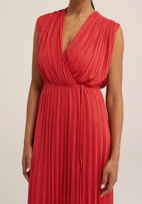 Röd plisserad klänning med djup V-ringning och korta ärmar. Mjuk tyg med insvängd midja och knytdetalj. Lätt texturerad yta.