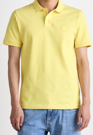 Muž v krátkém žlutém polo tričku se dvěma bílými knoflíky, doplněném světle modrými džínami.