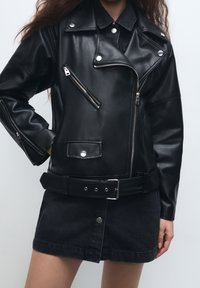 Donna che indossa una giacca da motociclista nera in pelle con zip e bottoni argentati, abbinata a una mini gonna nera con bottoni frontali e cintura.