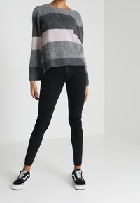 Grauer und rosa gestreifter, flauschiger Pullover mit weitem Ausschnitt, kombiniert mit schwarzen Skinny-Jeans und schwarzen Sneakern an einem Model.