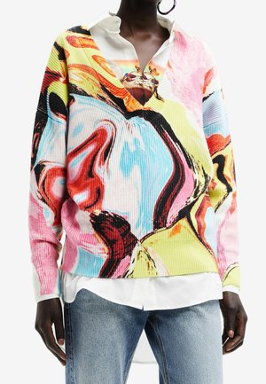 Osoba ubrana w sweter z abstrakcyjnym, wielokolorowym wzorem wirującym, nałożony na białą koszulę z kołnierzykiem i niebieskie dżinsy, widok na środek tułowia.