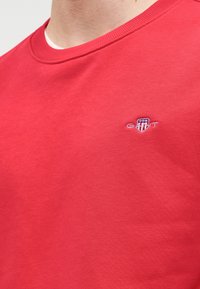 Sweat-shirt en coton rouge avec un col rond et des poignets côtelés. Présente un petit logo brodé sur la poitrine. Texture douce, design décontracté.