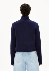 Veste zippée en polaire navy avec un col montant, un design court et des manches longues ; portée sur un pantalon en denim bleu clair avec des poches arrière.