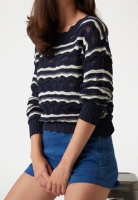 Marineblau-weiß gestreifter Strickpullover mit strukturiertem Muster, Rundhalsausschnitt und gerippten Bündchen. Kombiniert mit blauen Shorts, was einen lässigen Look zeigt.