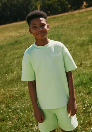 Junge lächelt, trägt hellgrüne Shorts und T-Shirt mit der Aufschrift "Palm Beach Florida" und steht in einem sonnigen, grasbewachsenen Park mit Bäumen im Hintergrund.