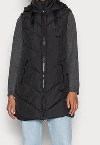 Schwarze Steppweste mit durchgehendem Reißverschluss, Chevron-Steppmuster, verstellbare Kapuze und zwei Reißverschlusstaschen. Über einem grauen Langarmshirt getragen.