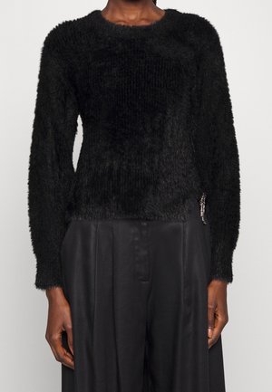 Personne portant un pull noir à manches longues duveteux associé à un pantalon ample en satin noir, les mains détendues le long du corps, sur un fond uni.