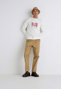White sweatshirt featuring a printed design, paired with tan trousers and brown shoes. Individual wears a light brown hat.