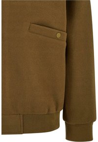Manche et côté de veste en laine marron avec une poche à rabat sécurisée par un bouton-pression doré et des détails côtelés au niveau du poignet et de l'ourlet.