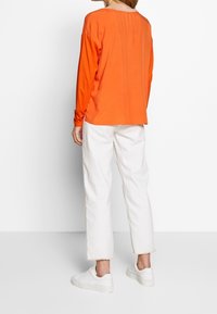 Person trägt ein orangefarbenes Langarmshirt, weiße gekürzte Hosen mit ausgefransten Säumen und weiße Turnschuhe, steht vor einem schlichten weißen Hintergrund.