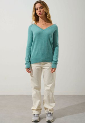 Maglione di lana azzurro chiaro con scollo a V, orlo a coste e maniche lunghe, abbinato a jeans avorio e sneaker grigie su uno sfondo neutro.
