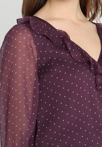 Lila durchsichtige Bluse mit tiefem V-Ausschnitt und Rüschensaum, versehen mit weißen Punkten und langen, durchscheinenden Ärmeln.