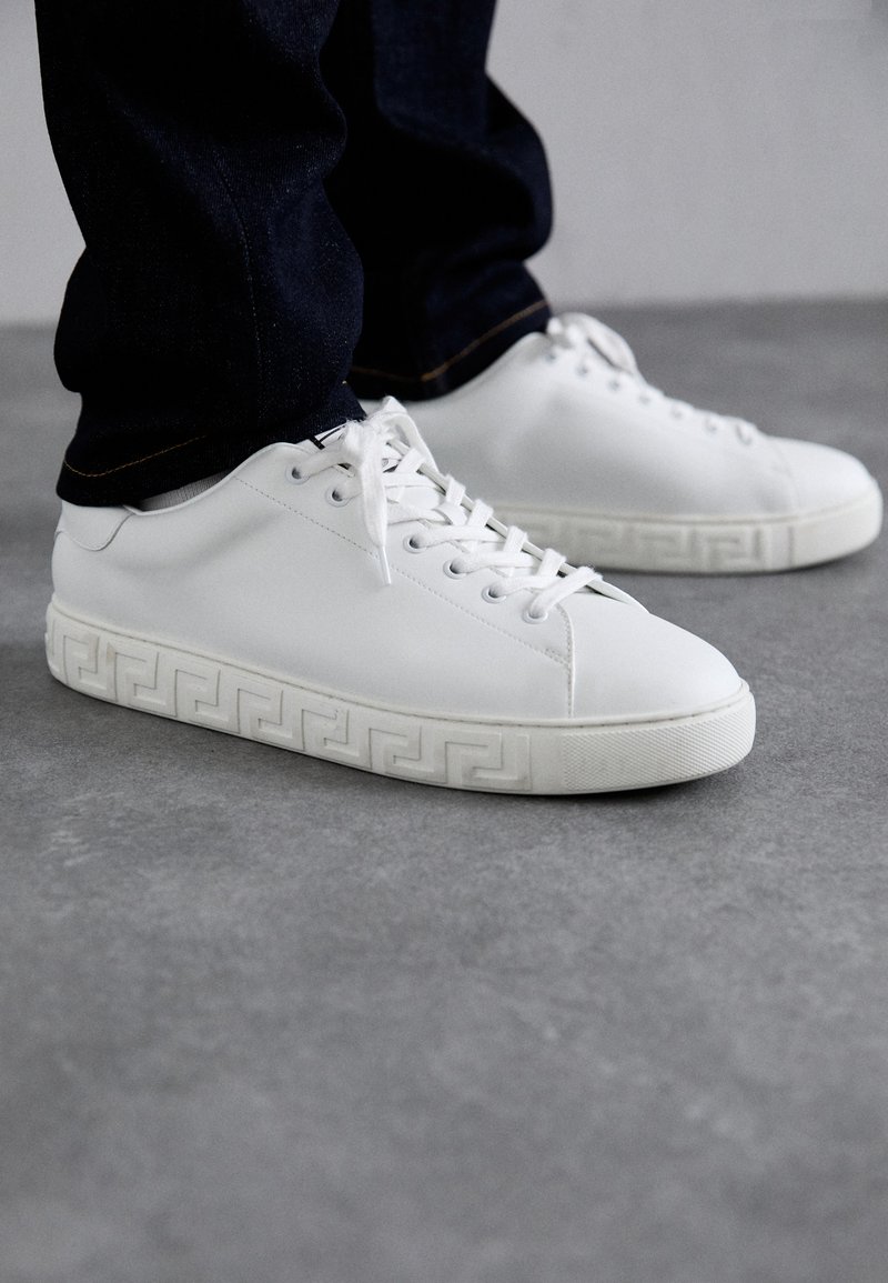 Witte leren sneakers met een gestructureerde zool met een Grieks sleutelpatroon, gecombineerd met donkere spijkerbroeken. Platte veters en een minimalistisch ontwerp.