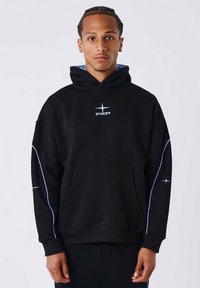 Jonge man met gevlochten haar, gekleed in een zwarte hoodie met blauwe piping en het PXP-logo, staand tegen een effen lichte achtergrond.