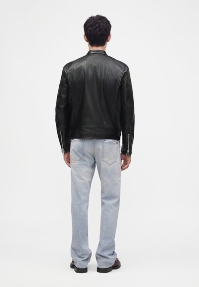 Homme portant une veste en cuir noire et un jean bleu clair usé, debout, tourné de dos sur un fond blanc uni.