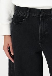 Zwarte denim jeans met een klassieke pasvorm, voorzien van een voorzak en een standaard metalen knoopsluiting. De textuur is glad met een lichte glans.