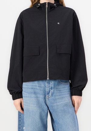 Sort lynlåsjakke med hætte, elastiske manchetter, klaplommer og lille "cK" logo på brystet, båret med lyseblå jeans.