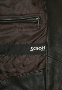 Brunt quiltet stoffinteriør med en glidelåslomme og svart stoffmerke med teksten "Schott N.Y.C." sydd innvendig i en svart skinnjakke.