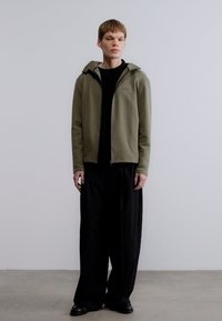 Ζακέτα ελιάς με φερμουάρ πάνω από μαύρο πουκάμισο· συνδυασμένη με φαρδιά μαύρη παντελόνα και μαύρα παπούτσια. Απλός σχεδιασμός και άνετη εφαρμογή.