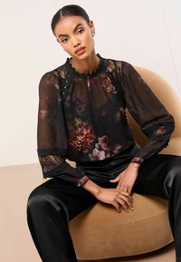 Čierna kvetinová blúzka z priesvitného materiálu, s čipkovými detailmi a vysokým golierom. Skombinovaná s čiernymi saténovými nohavicami. Sedí na okrúlej béžovej stoličke.