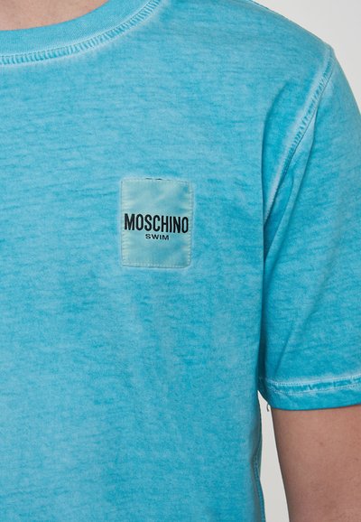 T-shirt in cotone blu chiaro con una toppa rettangolare con la scritta "MOSCHINO SWIM" in testo nero. Collo rotondo e maniche corte.