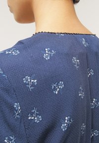 Gros plan du haut du dos d’une personne portant un vêtement bleu avec de petits motifs floraux blancs et une bordure perlée au niveau de l'encolure.