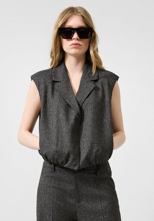 Frau trägt ärmellosen dunkelgrauen Blazer und passende Hose mit schwarzen übergroßen Sonnenbrillen vor einfachem Hintergrund.