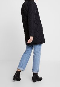 Černá prošívaná péřová bunda s dlouhými rukávy, standardním límcem a bočními kapsami, v kombinaci se světle modrými zahnulými džíny a černými kotníkovými botami.