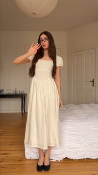 Γυναίκα με μακριά σκούρα μαλλιά και γυαλιά, που φοράει λευκό φόρεμα και μαύρα παπούτσια, στέκεται δίπλα σε ένα κρεβάτι σε ένα απλό υπνοδωμάτιο, σηκώνοντας το δεξί της χέρι.