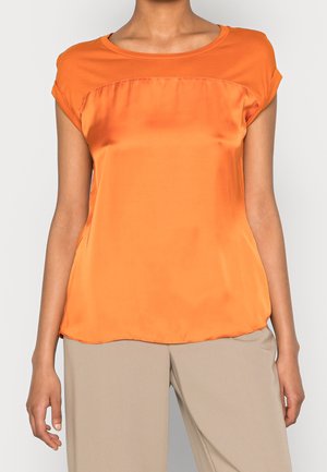 Ærmeløs orange satinbluse med rundet halsudskæring, båret sammen med beige løstsiddende bukser på en person, der står foran en ensfarvet baggrund.