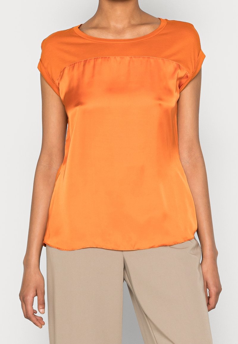 Blouse sans manches en satin orange avec un col arrondi portée avec un pantalon ample beige par une personne debout devant un fond uni.